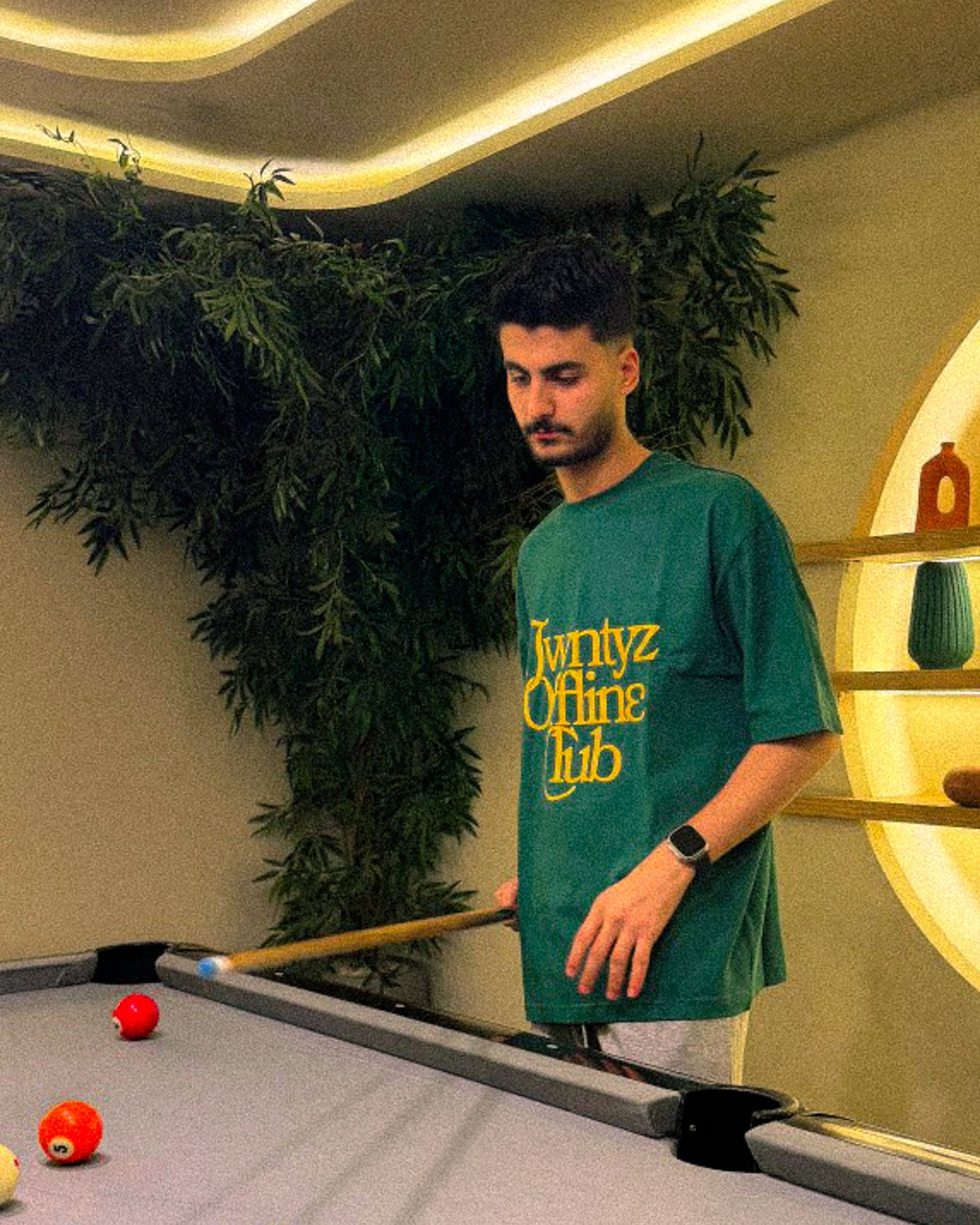 Man in a green t-shirt standing by a pool table with pool balls and a cue.