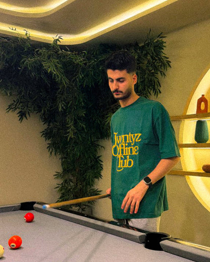 Man in a green t-shirt standing by a pool table with pool balls and a cue.